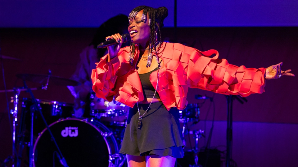 Poundo Gomis is performing on stage with a microphone in her right hand and her left arm extended outward. She is wearing a bright red textured jacket over a black outfit and has beaded braids. She is under purple and yellow stage lighting. A drum set is visible in the background.