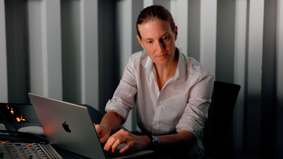 Dorien Herremans, wearing a white shirt, working on a MacBook in a dimly lit room with vertical, evenly spaced wall panels behind her.