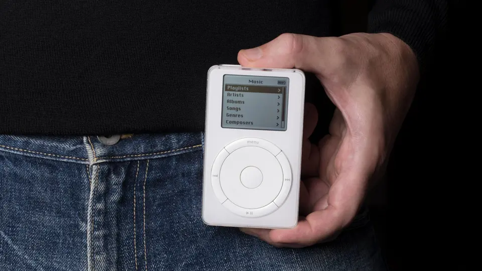 ジーンズと黒いセーターの前で左手に初代iPodを持つ人。デバイスの小さなモノクロ画面には、プレイリスト、アーティスト、アルバムなどのメニューオプションが表示され、その下に円形のクリックホイールを備える。